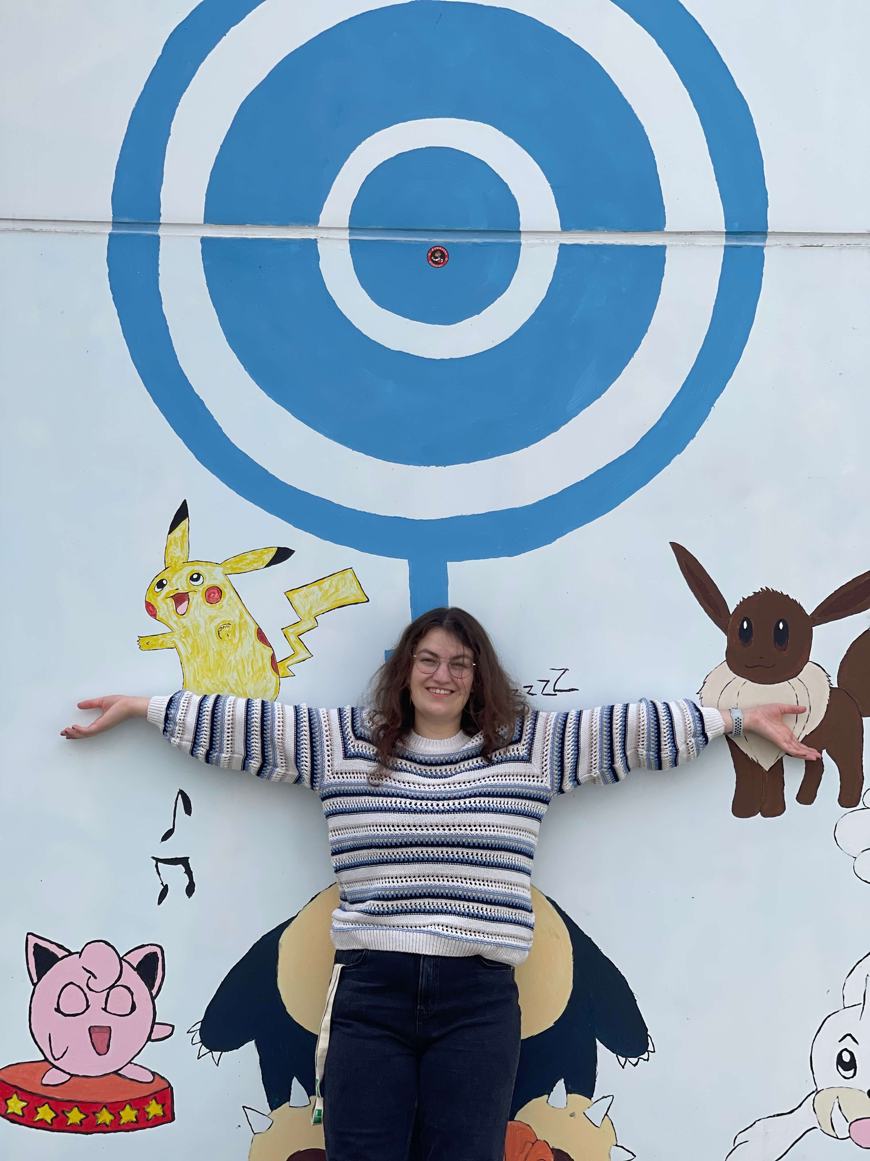 A student in a sweater poses in front of a Pokemon mural on a white wall