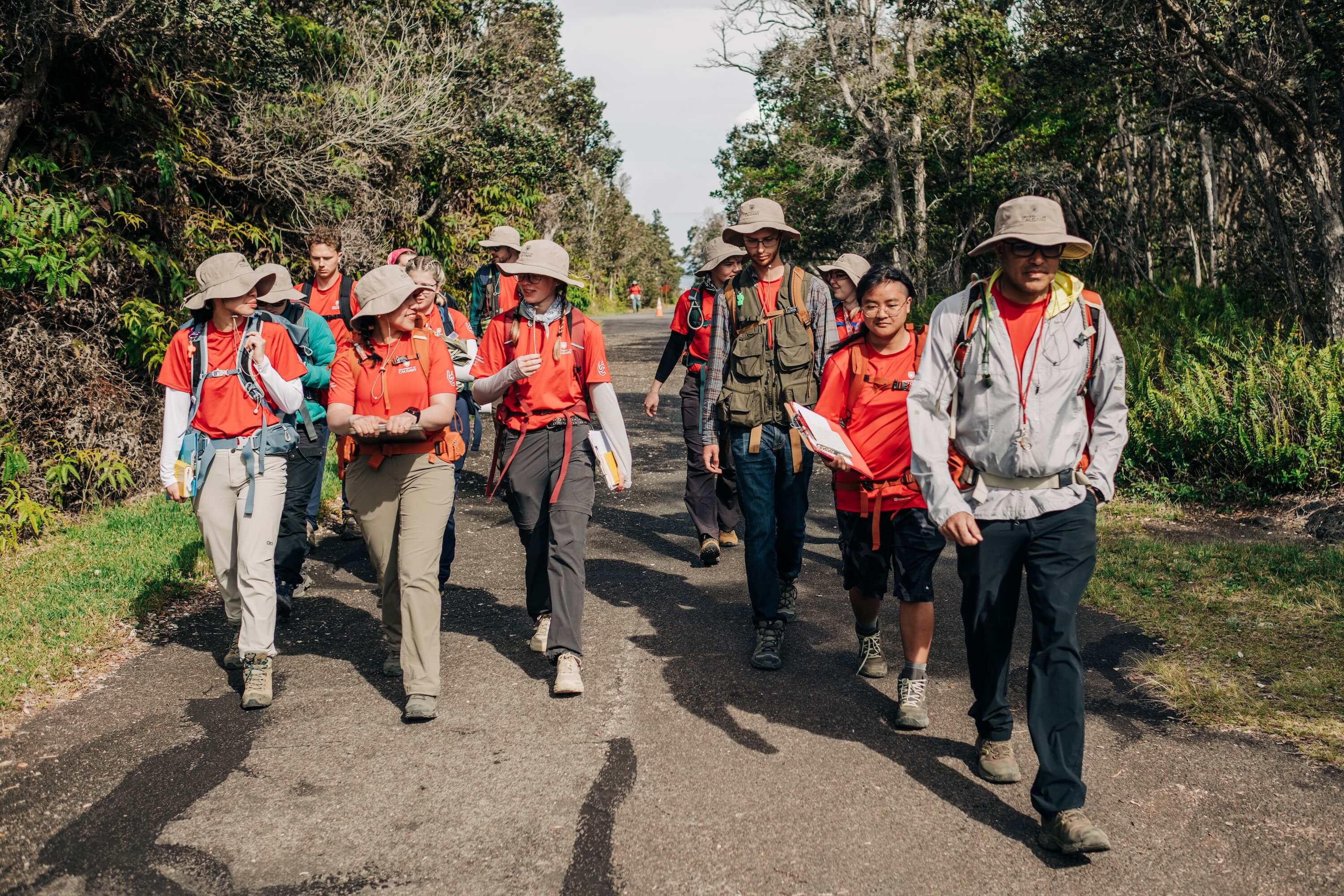A group of students in orange outfits walks along a path surrounded by plants