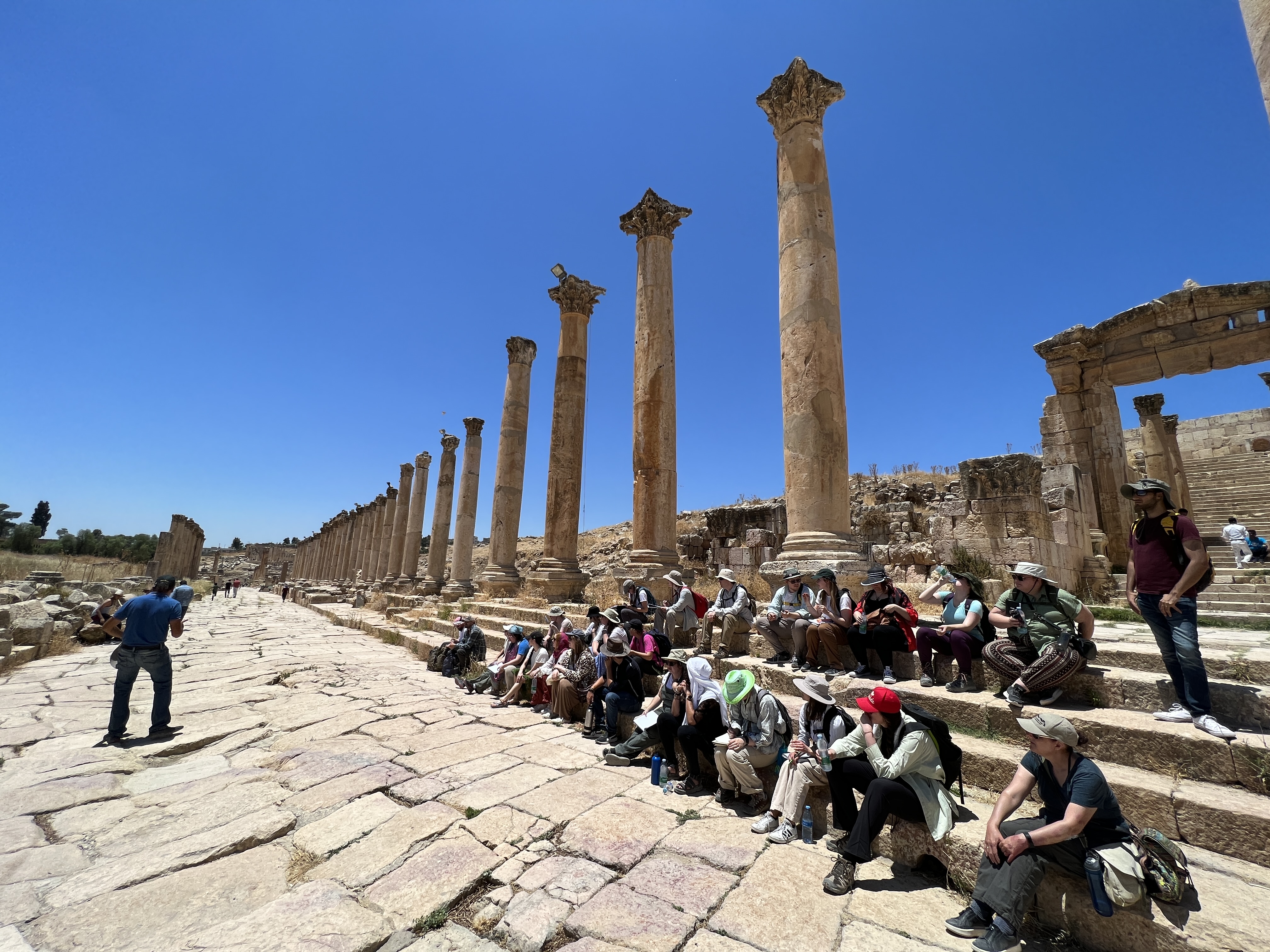 A group of travellers sit beneath tall stone pillars, listening to a guide speak.