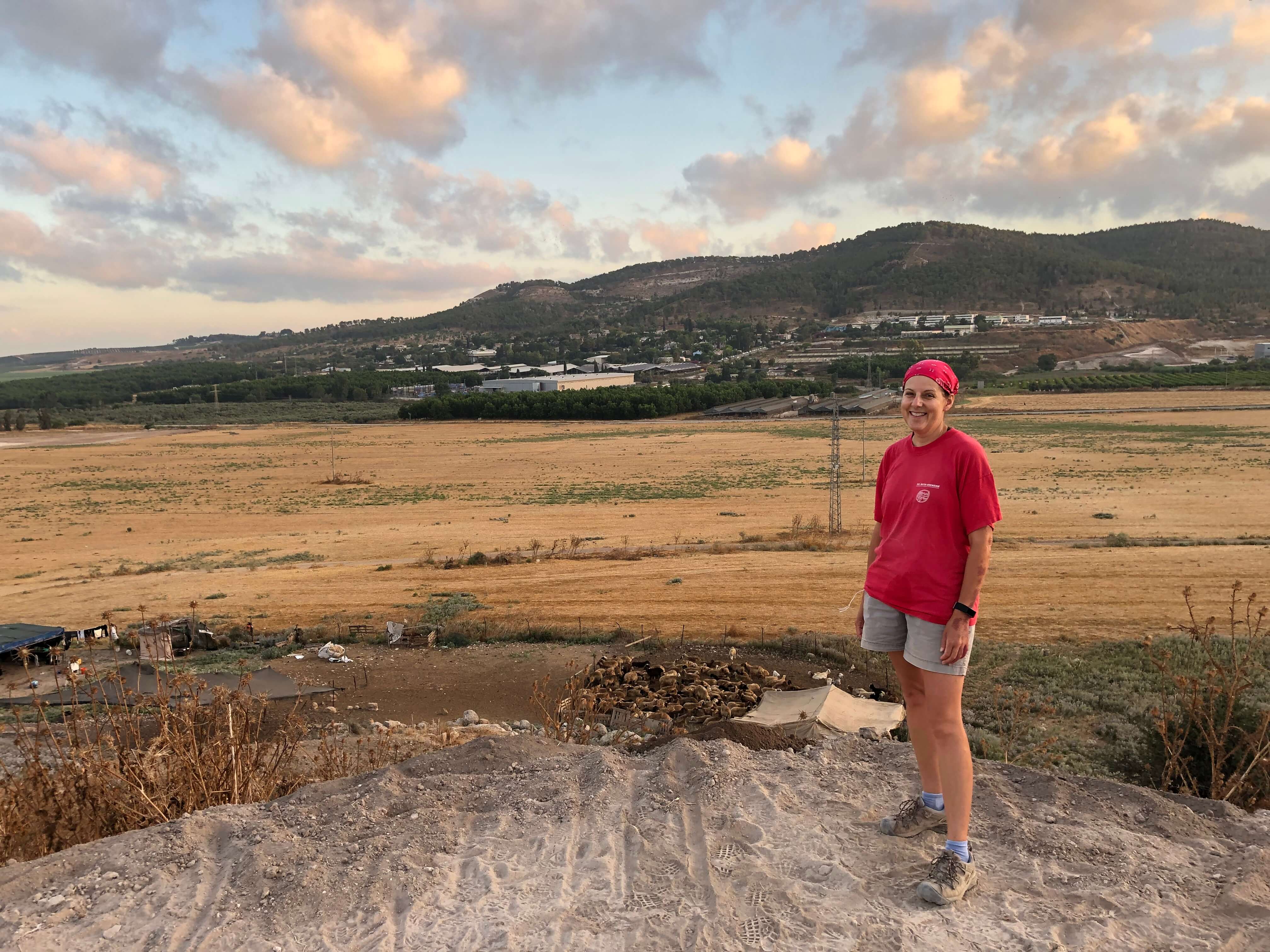 A person in a red shirt and shorts standing with a field and hill in the background